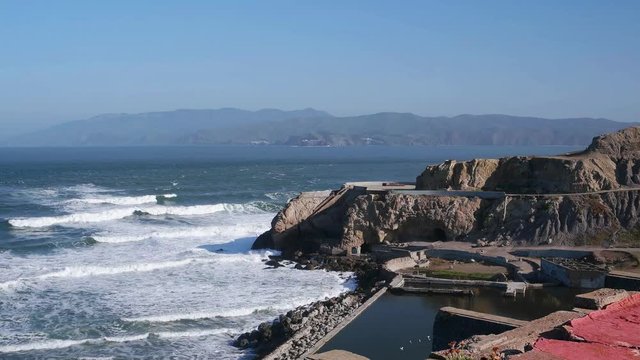 Time lapse of Sutro Baths on a sunny spring morning.  Surf rolls in from the Pacific.  Sea gulls bath on the remains of the swimming pool.  A large ship steams by.  Clear blue sky.