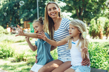Fototapeta premium Waist up portrait of happy pretty woman sitting with kids on bench in park. Small girls are holding lollypops and catching soap bubbles with hands. They are smiling and content