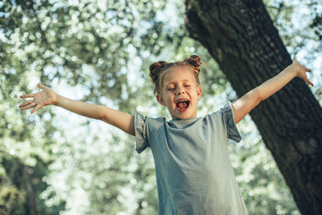 Hurray. Waist up portrait of cheerful girl standing at tree with hands stretched. She is screaming in delight with open mouth and closed eyes 
