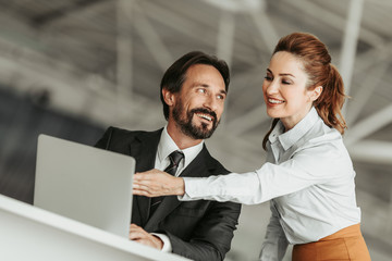 Low angle portrait of glad unshaven employer talking with happy secretary. She pointing at notebook computer indoor