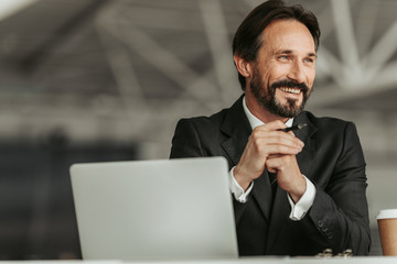 Portrait of smiling bearded businessman holding pen in arms while working with laptop
