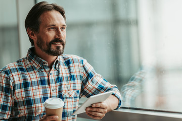 Portrait of smiling bearded male enjoying appetizing cup of coffee while using gadget indoor