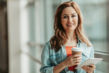 Portrait of happy girl tasting mug of hot liquid while typing in electronic tablet. Glad lady using appliance during rest concept