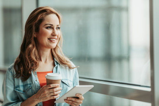 Portrait of smiling female drinking cup of coffee while holding digital device in hand. She looking at window