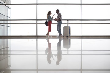 Full length side view happy lady gesticulating hands while speaking with glad male tourist in airport near baggage. Their reflection situating on white clear floor indoor