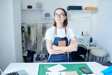 Portrait of young clothier standing in an apron and with her arms crossed in her workshop