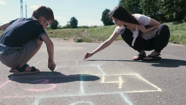 Mother and son drawing together hopscotch on the asphalt.