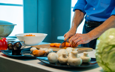 A man in a blue shirt, in the kitchen, on a wooden board cuts carrots