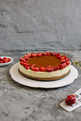 Cheese cake with strawberries and vanilla cream. Spoon of caramel and plate with strawberries is on the grey background
