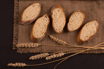 Pieces of sliced ​​baguette lie on a dark jute napkin.  Nearby lie a few spikelets of wheat