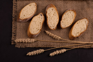 Pieces of sliced ​​baguette lie on a dark jute napkin.  Nearby lie a few spikelets of wheat