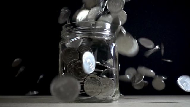Slow motion, a pile of coins falls into an empty glass jar on a black background.  Ukrainian coins fall into a jar.