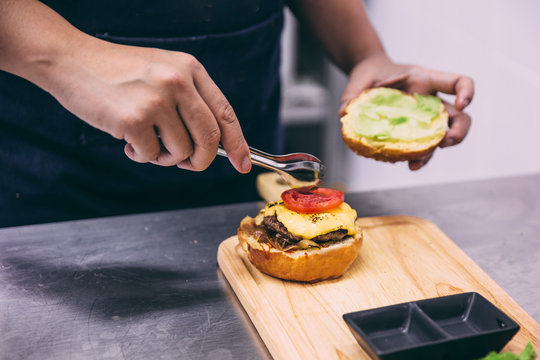 Chef add a sliced of tomato for making a beef cheeseburger on wooden plate.   