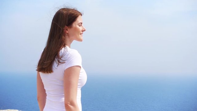 Smiling young woman in white t-shirt standing at edge of cliff on sunny day in slow motion. Carefree female looking at breathtaking view of blue Mediterranean sea. Concept of freedom and inspiration