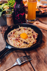 Fettuccine Carbonara yolk bacon served in black pan on wooden table with French fries in the background.