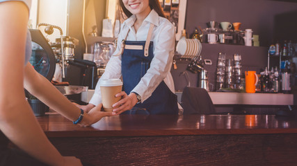 Young beautiful Asian woman barista wear blue apron holding hot coffee cup served to customer at bar counter in coffee shop with smile face.Concept of cafe and coffee shop small business.Vintage tone