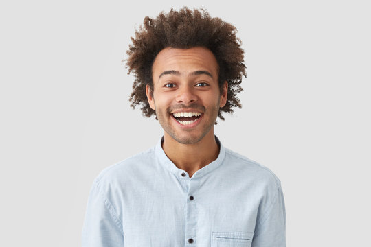 Black overjoyed curly male with shining smile, shows white teeth, being glad to recieve praise for diligent work, stands indoor over studio wall. Cheerful African American man expresses positiveness