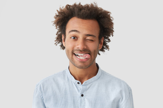 Positive attractive African American male with positive expression, shows tongue, has happy expression, stands against white wall, has crisp hair, have fun together with friends. Funny hipster