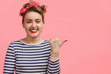 People, advertisement and emotions concept. Beautiful cute young woman with make up, red lips, points aside with thumb, has shining smile, shows blank space for your promotional content or text