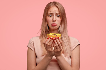 Discontent blonde female holds sweet doughnut, looks with temptation, wants to eat, but keeps to diet, dressed in casual t shirt, isolated over pink background. People and junk food concept.