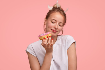 People and temptation concept. Attractive young European female looks at sweet doughnut, going to eat for breakfast, fonds of junk food, dressed casually, isolated over pink studio background