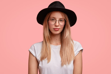 Portrait of stylish European female student wears spectacles and fashionable black hat, has serious expression, stands against pink background, talks with groupmates about exam. Youth concept