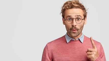 Emotive Caucasian male with shocked expression, looks in bewilderment at camera, raises fore...
