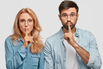 Portrait of mute pretty young female and male makes hush sign, ask to be quiet, keep fore fingers on lips, stand closely, isolated over white background. People, conspiracy and secrecy concept