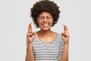 Portrait of adorable young female with positive smile, has Afro hairstyle and dark skin, crosses fingers as believes in good luck, dressed in striped sweater, isolated over white studio background