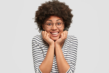 Fototapeta na wymiar Optimistic curly African American female has toothy smile, keeps hands under chin, listens pleasant story from friend, wears casual clothes and round glasses, isolated over white background.