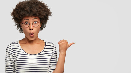 Emotive lovely mixed race female with stupefied expression has amazed look, opens mouth, points with thumb at blank space for your advertisement or promotional content. Omg, just look there!