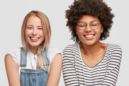 Two cute diverse women laugh positively, have broad smiles, hear funny anecdote from friend, spend free time together after having classes, express happiness, isolated over white studio wall