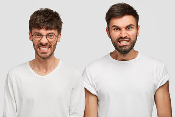 Studio shot of angry furious bearded men colleagues clench teeth in annoyance, feel irritation as recieve much work and duties from boss, dressed in casual white t shirts. Negative human emotions