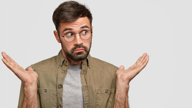 Confused attractive unshaven man shrugs shoulders, looks with hesitation aside, purses lips in bewilderment, has clueless expression, isolated over white background with free space for advertisement