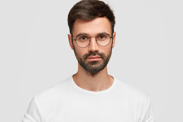 Fototapeta premium Headshot of handsome male freelancer with appealing look, has dark beard and mustache, looks directly at camera with serious look, wears white casual clothes. Monochrome. Facial expressions.