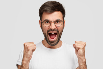 Angry bearded guy with mad expression, raises eyebrows with anger, scream sloudly, wears casual white t shirt, expresses annoyance, feels crazy, isolated over studio wall. Don`t make noise, please!