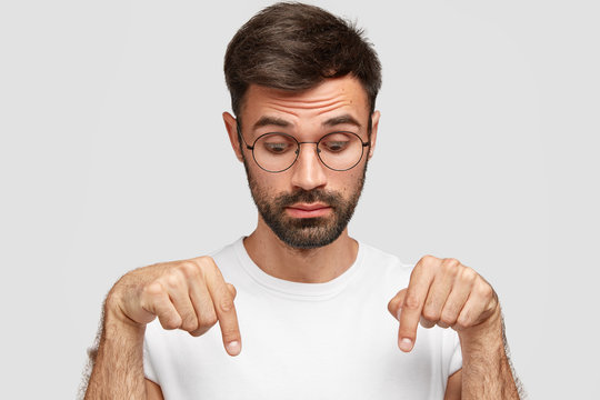 Headshot of handsome bearded male points down with surprised look, notices something on floor, wears glasses, dressed in casual t shirt, isolated over white background. People and astonishment