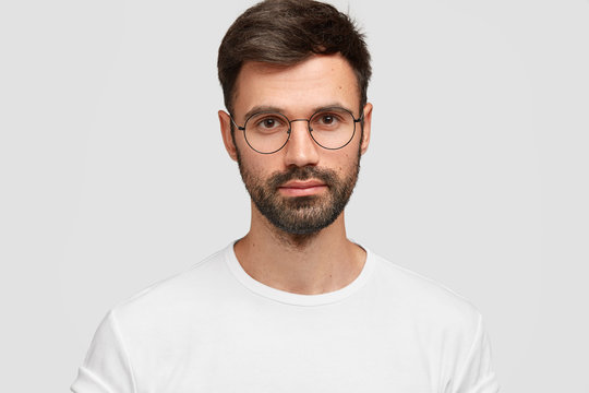 Headshot of handsome male freelancer with appealing look, has dark beard and mustache, looks directly at camera with serious look, wears white casual clothes. Monochrome. Facial expressions.
