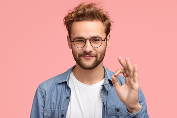 Portrait of handsome man has stubble, makes ok sign, agrees or likes something has joyful...