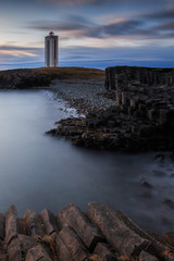 Fototapeta premium Leuchtturm im letzten Abendlicht. Im Vordergrund Basaltgestein. Im Norden Islands