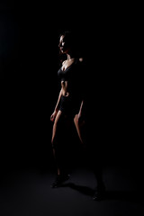 A sports girl in black shorts and a topic is standing on a black isolated background. Female portrait in a low key.