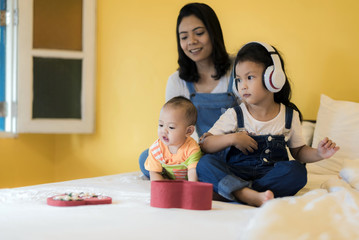 Babysitting playing with three foolish playful kids carrying on the bed; widow young woman and child are enjoying funny time indoor in modern yellow bed room.