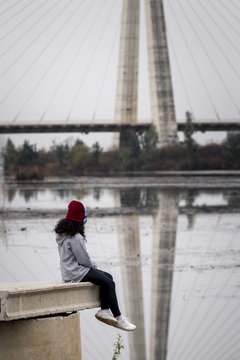 Niña escuchando música sentada al lado del río junto al puente de la ciudad