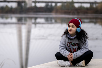 Niña escuchando música sentada al lado del río junto al puente de la ciudad