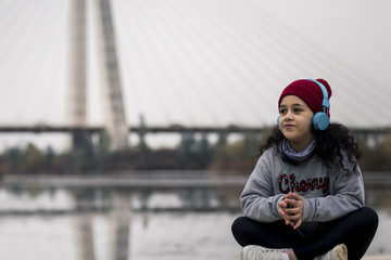 Niña escuchando música sentada al lado del río junto al puente de la ciudad