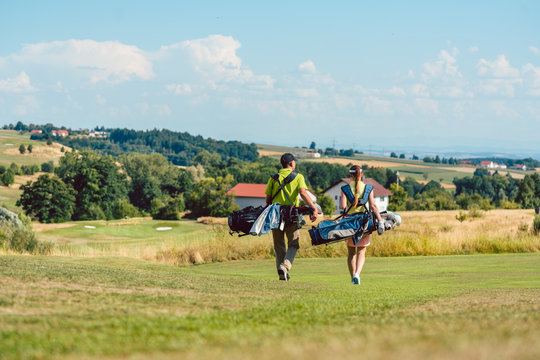 Full length rear view of a couple carrying professional golf bags, while walking on green field towards the golf course in a sunny day of summer - Powered by Adobe