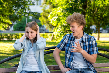 A young guy screams at a girl. Summer in park on a bench. The girl covered her ears with her hands. The concept of aggression is a problem in relations. Conflict in the family.