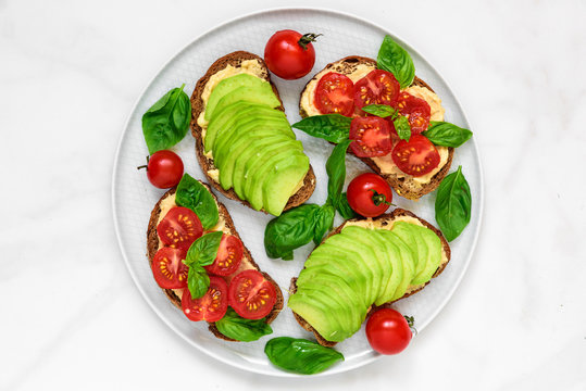 healthy breakfast toasts with avocado, tomatoes, hummus and basil over white marble table
