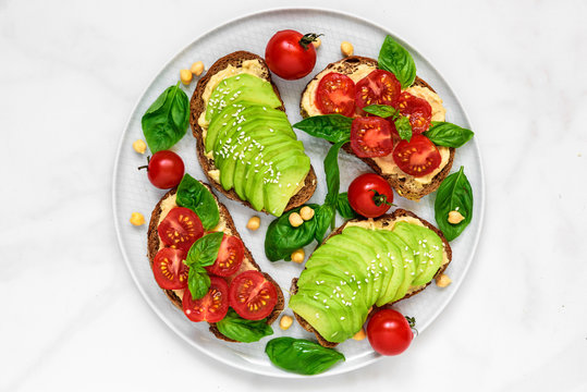 Avocado and tomatoes toasts with hummus, sesame and basil in a plate over white marble background. vegan food
