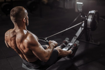 close up back view image. well-built guy is warming up with rowing machine. full length photo